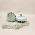 Two massage brushes showing the ergonomic handle displayed on stone with a beige background